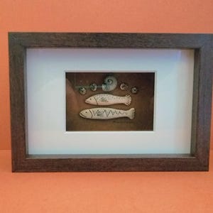 May include: Framed artwork featuring two stylized fish and a nautilus shell. The fish are white with black detailing, set against a brown background. The frame is a dark brown color, and the artwork is matted with a white border.