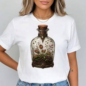 May include: A white short-sleeved t-shirt featuring a central graphic of a clear glass apothecary bottle with a cork stopper. Inside the bottle is a detailed illustration of a green and red Venus flytrap plant growing in dark soil.