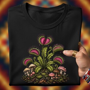 May include: A black t-shirt featuring a graphic illustration of a green Venus flytrap plant with vibrant pink interiors and spiky edges. The plant is surrounded by a cluster of small, earthy-toned mushrooms and mossy ground details.