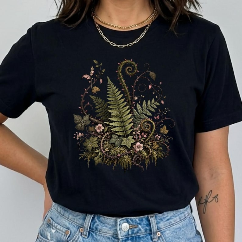 May include: A black short-sleeved t-shirt featuring a central graphic print of green ferns, thorny vines, and small pink flowers. The botanical illustration displays detailed foliage, curled fronds, and mossy textures centred on the chest area.