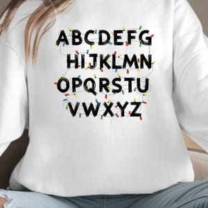 May include: White sweatshirt with the alphabet in black letters, each letter is decorated with colorful string lights. The letters are arranged in rows, with the letters A through Z displayed.