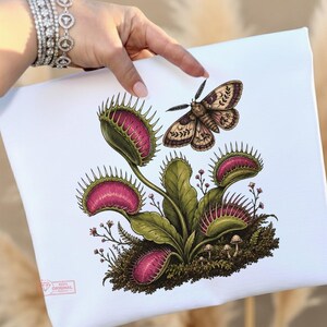 May include: A white canvas featuring a detailed illustration of a green Venus flytrap with vibrant pink interiors, surrounded by small mushrooms and moss. A moth with patterned wings is positioned above the plant. A small logo is in the corner.
