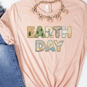 May include: A light pink t-shirt with the words "Earth Day" in a colorful, stitched design. The letters are in shades of green, blue, and tan, with a tree and sun graphic. A gold-toned necklace with floral accents is draped on the shirt.