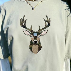 以下が含まれることがあります： 鹿の頭のデザインが施された、薄い緑色のTシャツ。鹿の頭は、さまざまな迷彩柄、花柄、青い正方形で構成されています。Tシャツは半袖で、クルーネックです。