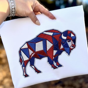 Puede incluir: Lienzo blanco con un diseño geométrico de un bisonte en rojo, blanco y azul. El bisonte está bordado con un patrón poligonal, creando una estética moderna y patriótica. La obra de arte está sostenida por una mano.