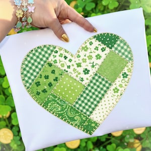 Puede incluir: Un lienzo blanco con un diseño de corazón de patchwork en varios tonos de verde. El corazón está compuesto por cuadrados con patrones como lunares, cuadros y estampados florales, cosidos con un hilo contrastante.