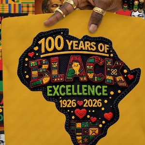 Op de afbeelding: Een gele t-shirt met een zwart geborduurde kaart van Afrika. De kaart is gevuld met kleurrijke patronen en de tekst "100 Years of Black Excellence 1926-2026" in goud en groen. Het ontwerp is gedetailleerd en levendig.