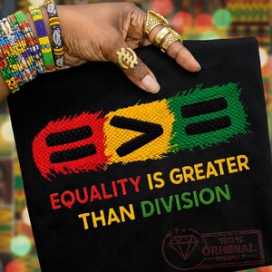 Könnte beinhalten: Schwarzes T-Shirt mit dem Text "EQUALITY IS GREATER THAN DIVISION" in Gelb, Grün und Rot. Das Design umfasst ein Gleichheitszeichen, ein Größer-als-Zeichen und ein weiteres Gleichheitszeichen. Das Shirt hat auch ein Diamant-Logo und die Worte "100% ORIGINAL".