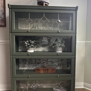 May include: A dark green wooden bookcase with glass doors and shelves. The bookcase has a decorative wallpaper design on the back of the shelves. The bookcase is decorated with a small potted plant, a book, and a geometric metal sculpture.