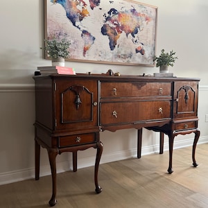May include: A dark wood antique buffet with ornate details, featuring drawers and cabinet doors. The buffet has decorative hardware and stands on curved legs. A world map art piece hangs above the buffet, with small plants on top.