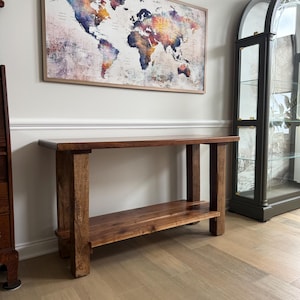 May include: A rustic, wooden console table with a lower shelf. The table is made of dark brown wood and has a rectangular top. A colorful world map in a wooden frame hangs on the wall above the table. A glass cabinet is to the right.