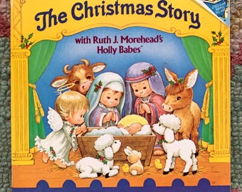 May include: A children's book titled "The Christmas Story" with illustrations of the Nativity scene. The cover features a yellow border with the title in blue script, and images of Mary, Joseph, baby Jesus, animals, and angels.