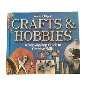 May include: A blue book with the title "Crafts & Hobbies: A Step-by-Step Guide to Creative Skills" by Reader's Digest. The cover features various craft tools and supplies, including a leather punch, a pair of pliers, a small wooden mallet, a paintbrush, and a small ceramic teapot.