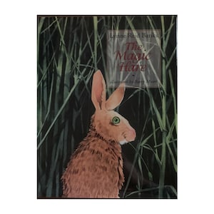 May include: The cover of a children's book titled "The Magic Hare" by Lynne Reid Banks, illustrated by Barry Moser. The cover features a brown hare with green eyes standing in tall green grass.