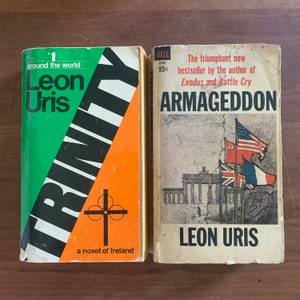 May include: Two vintage paperback books by Leon Uris. The book on the left is titled "Trinity" with a green and orange cover. The book on the right is titled "Armageddon" with a cover featuring flags and a building.
