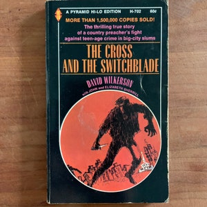 May include: A black and red book cover with the title "The Cross and the Switchblade" by David Wilkerson. The cover features a black and white illustration of a person holding a knife in a city setting.