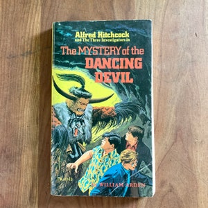 May include: A vintage paperback book cover featuring a scary illustration of a monster with horns and a wolf. The book title is "The Mystery of the Dancing Devil" by Alfred Hitchcock and The Three Investigators. The book is published by William Arden.