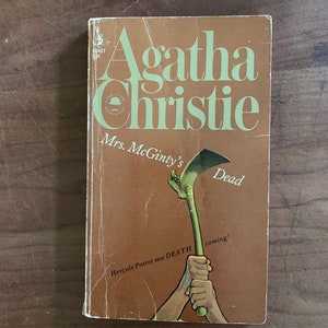 May include: A brown book cover with the title "Mrs. McGinty's Dead" by Agatha Christie. The cover features an illustration of a hand holding a large green sickle.