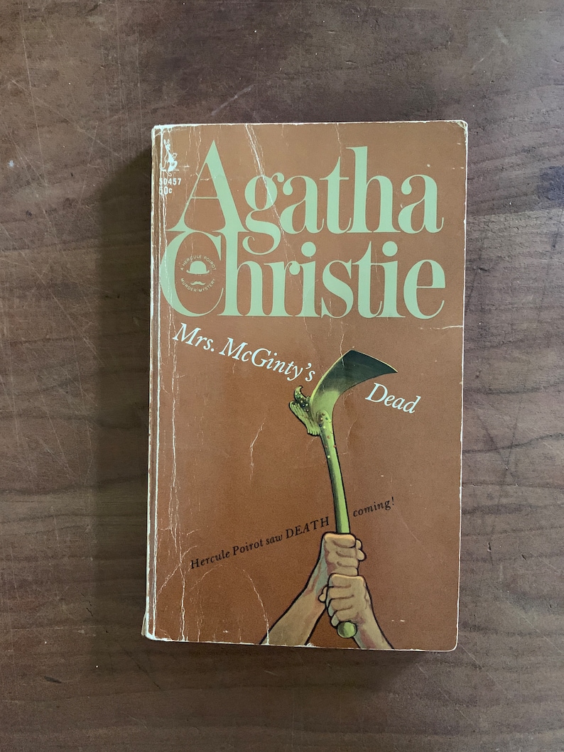May include: A brown book cover with the title "Mrs. McGinty's Dead" by Agatha Christie. The cover features an illustration of a hand holding a large green sickle.