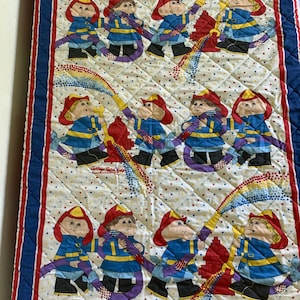 May include: A white quilted twin-sized bedspread with a red, blue, and yellow pattern. The pattern features a repeating design of Cabbage Patch Kids dressed as firefighters holding hoses and a fire hydrant. The text "Cabbage Patch Kids" is printed in the center of the bedspread.