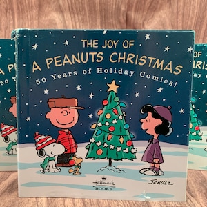 May include: A stack of "The Joy of a Peanuts Christmas" books. The cover features Charlie Brown, Snoopy, and Lucy standing in the snow near a decorated Christmas tree. The book's title is displayed with the text "50 Years of Holiday Comics!"