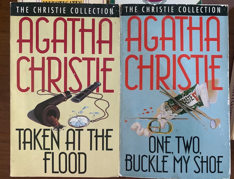 May include: Two paperback books from The Christie Collection featuring Agatha Christie novels. The book on the left is titled "Taken at the Flood" and features a pipe and a pocket watch on a yellow background. The book on the right is titled "One, Two, Buckle My Shoe" and features a blue background with a tube of toothpaste, a toothbrush, and a dental mirror.