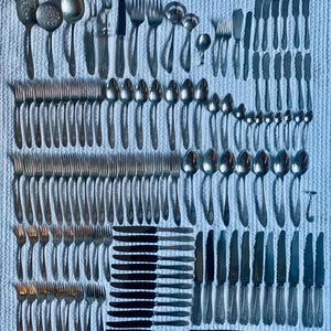 May include: An assortment of vintage silverware, including forks, knives, spoons, and serving utensils, arranged on a textured white surface. The silverware is made of a shiny, silver-colored metal, and the arrangement showcases a variety of designs and sizes.
