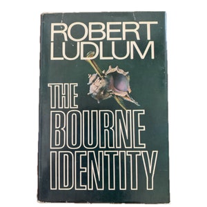 May include: A green book cover with the title "The Bourne Identity" in white letters. The author's name, "Robert Ludlum", is printed above the title in white letters. A seashell with a sharp object protruding from it is pictured on the cover.