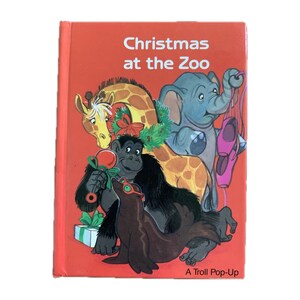 May include: A children's book titled "Christmas at the Zoo" with a red cover. The cover features cartoon illustrations of a giraffe, elephant, gorilla, and seal, all decorated for Christmas. The book is a Troll Pop-Up.