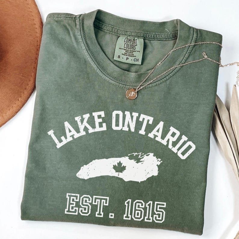 May include: An olive green short-sleeved t-shirt featuring white screen-printed text that reads LAKE ONTARIO and EST. 1615, with a white graphic of the lake containing a small maple leaf. The inner neck label reads COMFORT COLORS.