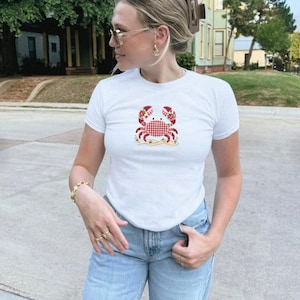 Peut inclure: T-shirt blanc à manches courtes avec un motif de crabe. Le crabe a un corps à carreaux rouges et blancs, des détails floraux rouges et est posé sur un fond de plage de sable. Le t-shirt est en matière souple, adapté au port décontracté.