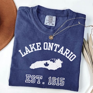 May include: A navy blue short-sleeved t-shirt with a distressed white graphic featuring the text LAKE ONTARIO, a map silhouette with a maple leaf, and EST. 1615. The shirt has a crew neckline and a Comfort Colors brand label inside the collar.