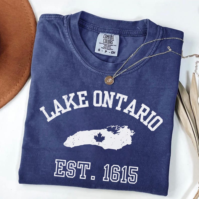 May include: A navy blue short-sleeved t-shirt with a distressed white graphic featuring the text LAKE ONTARIO, a map silhouette with a maple leaf, and EST. 1615. The shirt has a crew neckline and a Comfort Colors brand label inside the collar.