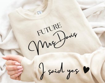 May include: A beige sweatshirt with the text "FUTURE Mrs. Davis" in black script across the chest. The sleeve displays "I said yes" with a black heart. The sweatshirt is folded on a white surface.