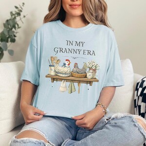 May include: Light blue t-shirt with the text "IN MY GRANNY ERA" above a graphic of a shelf with chickens, kitchen utensils, and flowers. The shirt is a soft, comfortable material.