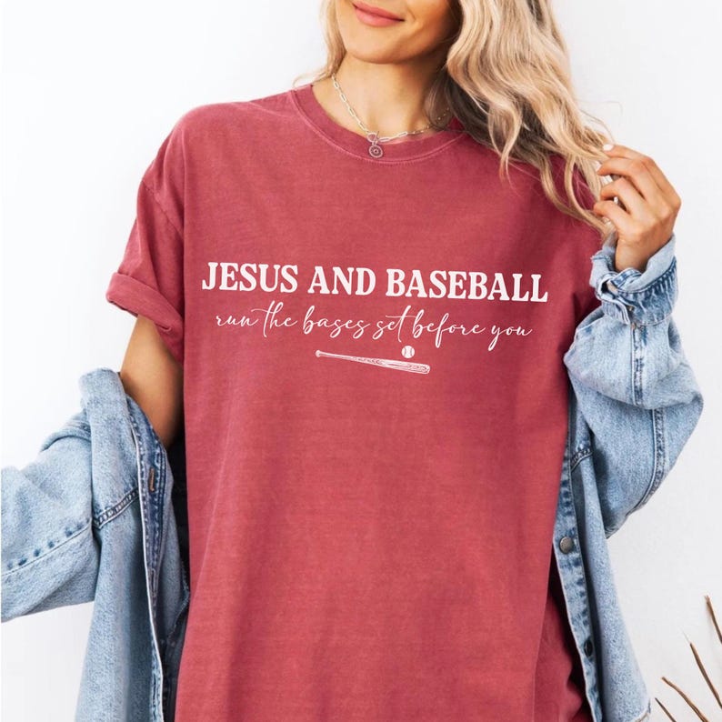 May include: A maroon t-shirt with the text "JESUS AND BASEBALL run the bases set before you" in white. A baseball and bat graphic is below the text. The shirt has rolled-up sleeves and is paired with a denim jacket.