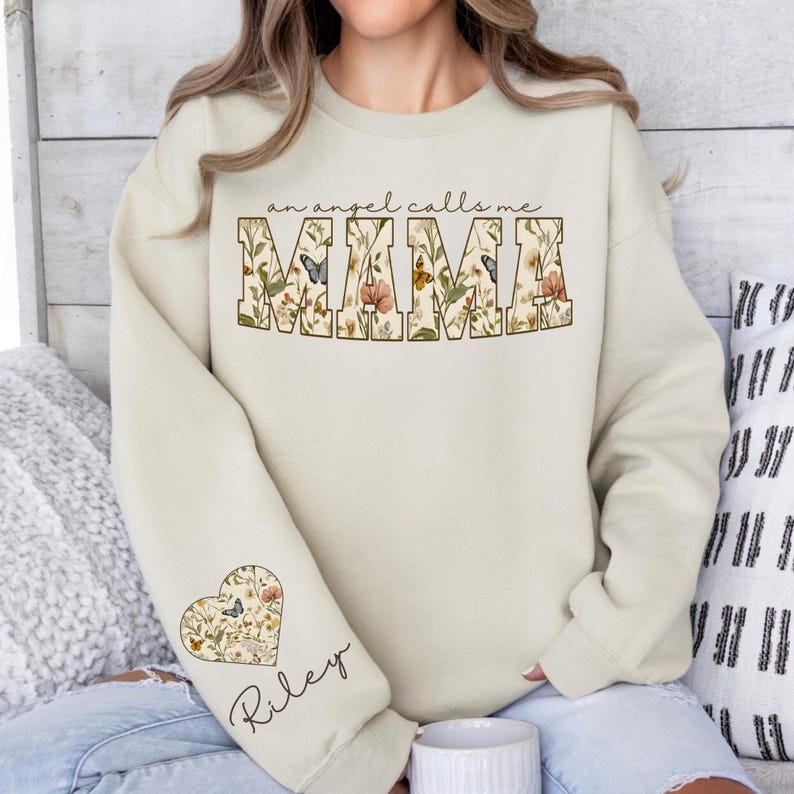 Pode incluir: Camisola creme com o texto "an angel calls me MAMA" em um design floral. A manga esquerda apresenta um design floral em forma de cora&ccedil;&atilde;o com o nome "Riley" escrito abaixo.