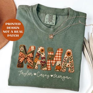 Puede incluir: Una camiseta verde salvia con la palabra MAMA impresa en estilo patchwork con patrones florales, de cuadros y de calabazas. Debajo de la palabra, los nombres Taylor, Casey y Morgan están impresos en letra cursiva blanca con corazones.