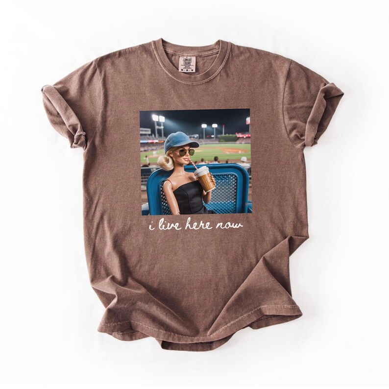 Pu&ograve; includere: T-shirt marrone con una grafica di una bambola a una partita di baseball, che tiene in mano una bevanda. La bambola indossa occhiali da sole e un berretto blu. Il testo "i live here now" &egrave; stampato sotto.
