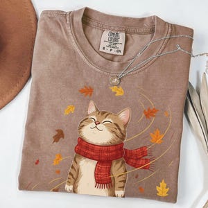 May include: A brown Comfort Colors t-shirt featuring a smiling cartoon cat wearing a red scarf, surrounded by falling autumn leaves. A silver necklace with a pendant rests on the shirt. The shirt is size S-P-CH.