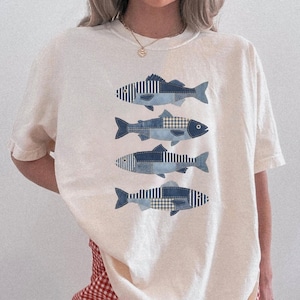 Könnte beinhalten: Cremefarbenes T-Shirt mit vier stilisierten Fischmotiven. Jeder Fisch ist ein Patchwork aus blauem Denim-Stoff mit Streifen- und Karomustern. Die Fische sind vertikal auf dem T-Shirt angeordnet.