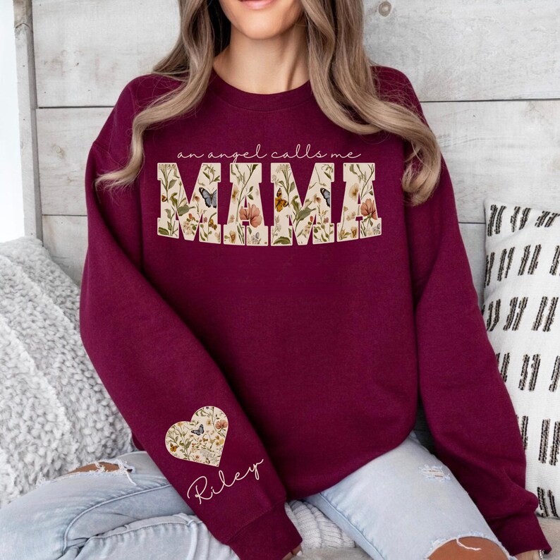 Pode incluir: Um moletom bord&ocirc; com o texto "an angel calls me MAMA" em um design floral. A manga apresenta um design floral em forma de cora&ccedil;&atilde;o com o nome "Riley" em script. &Eacute; uma pe&ccedil;a de roupa casual e confort&aacute;vel.