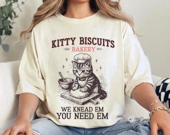 Può includere: T-shirt color crema con la scritta "KITTY BISCUITS BAKERY" sopra un'illustrazione di un gatto con cappello e grembiule da chef, che impasta. Il testo "WE KNEAD EM YOU NEED EM" è sotto l'illustrazione.