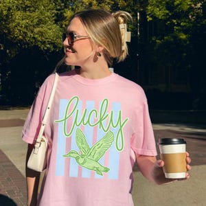 May include: Pink t-shirt with the word "Lucky" in green script, a green duck graphic, and light blue vertical stripes. The shirt is being worn by a person holding a coffee cup.