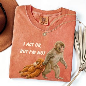 May include: Coral-colored t-shirt with the text "I ACT OK, BUT I'M NOT." The shirt features a graphic of two monkeys. One is lying down, and the other is walking. A gold necklace with a pendant is on the shirt.