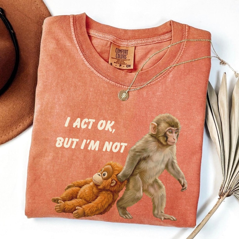 May include: Coral-colored t-shirt with the text "I ACT OK, BUT I'M NOT." The shirt features a graphic of two monkeys. One is lying down, and the other is walking. A gold necklace with a pendant is on the shirt.