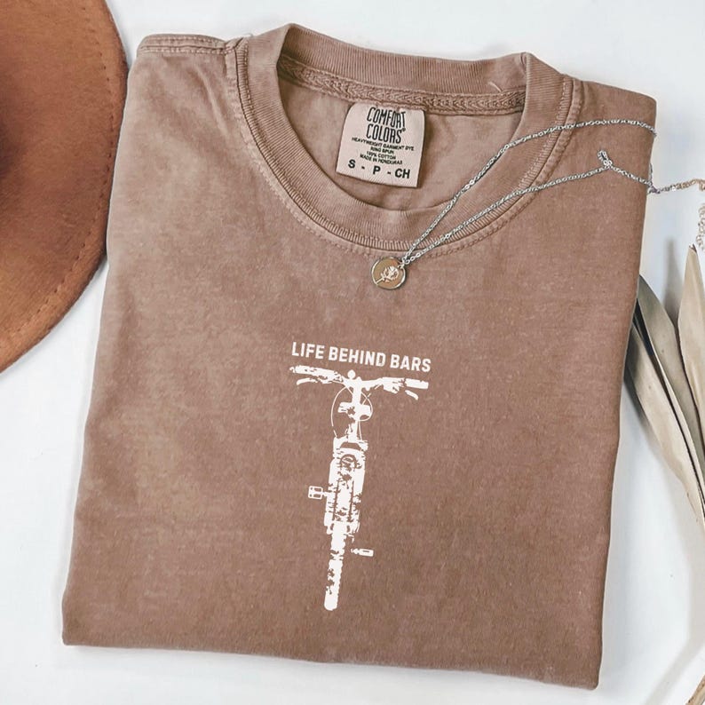 May include: A brown Comfort Colors t-shirt with the text "LIFE BEHIND BARS" and a white bicycle graphic. A silver necklace with a round pendant is on top of the shirt. The shirt is size S-P-CH.