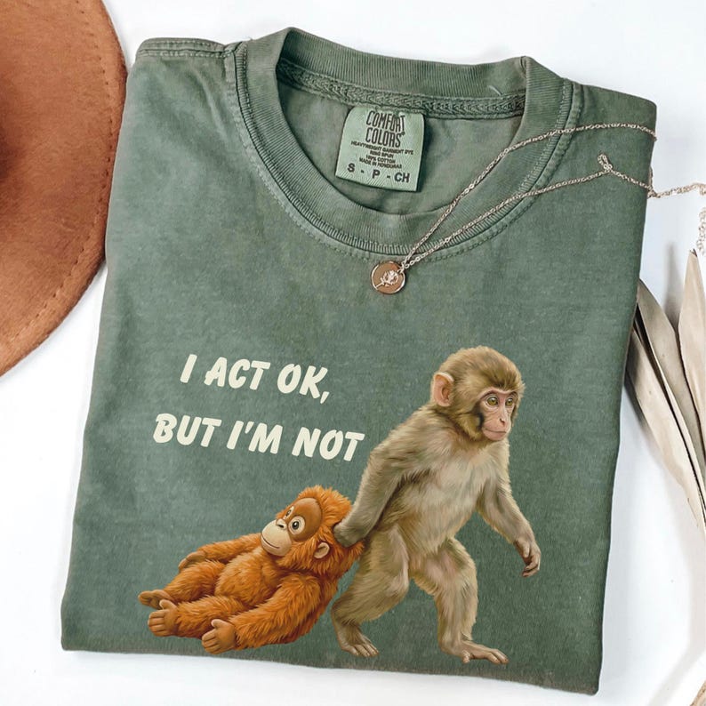 May include: Olive green Comfort Colors t-shirt with the text "I ACT OK, BUT I'M NOT" and a graphic of two monkeys. A silver necklace with a small pendant is on the shirt. The shirt is folded on a white surface.