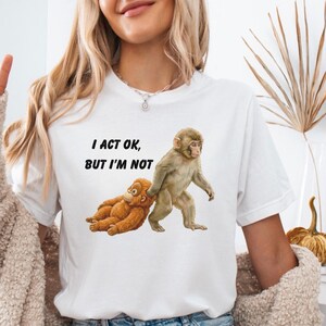 May include: White t-shirt with the text "I ACT OK, BUT I'M NOT" and a graphic of two monkeys. One monkey is standing, pulling the other monkey, who is lying down. The monkeys are brown and tan.