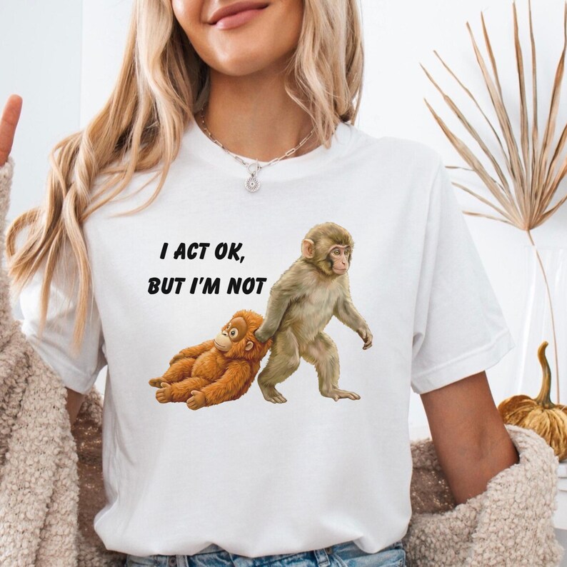 May include: White t-shirt with the text "I ACT OK, BUT I'M NOT" and a graphic of two monkeys. One monkey is standing, pulling the other monkey, who is lying down. The monkeys are brown and tan.
